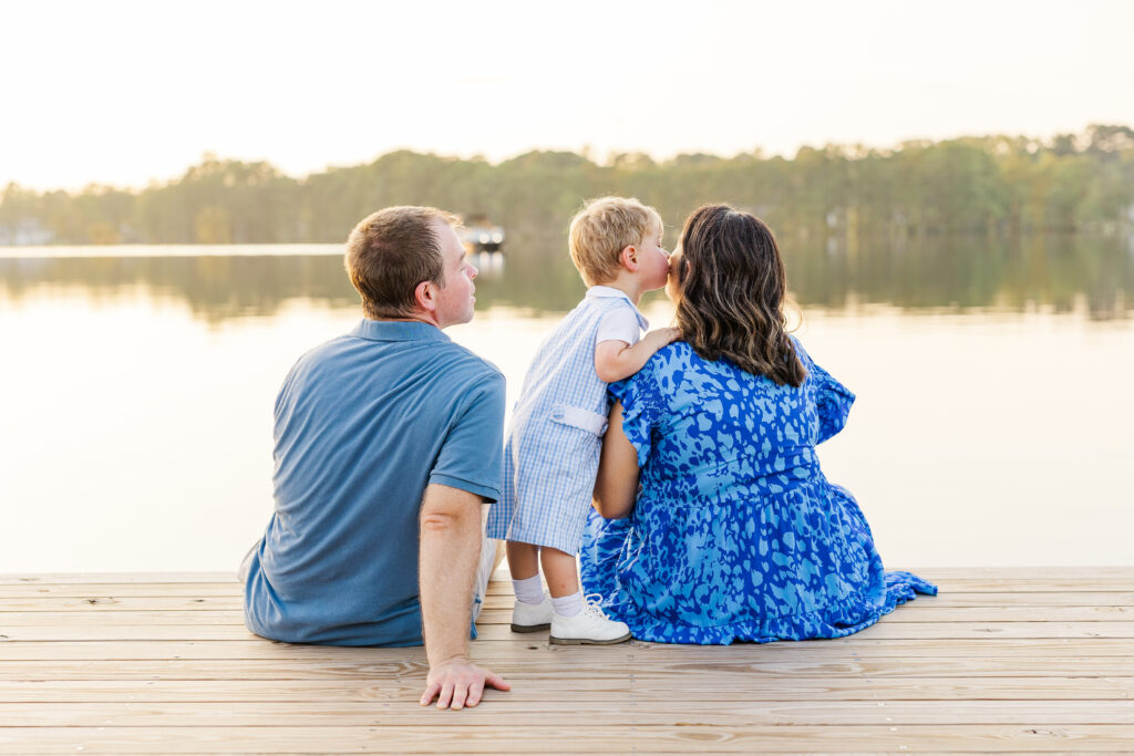 Son kissing mother on cheek next to dad sitting on dock on lake during Clayton Family photography session 