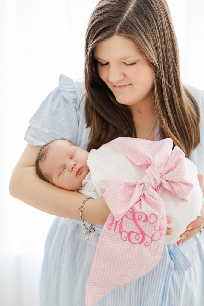 Mother holding her sleeping newborn wrapped in a pink monogrammed bow blanket during an Apex newborn photographer session.