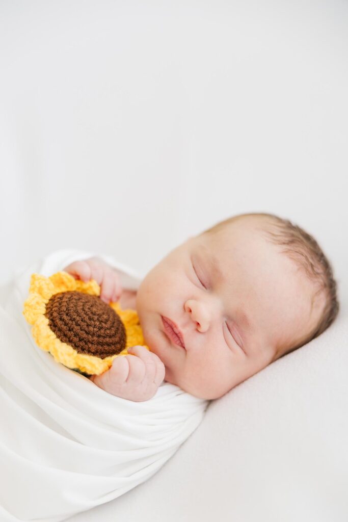 Sleeping newborn girl wrapped in white holding a sunflower prop, photographed by an Apex newborn photographer in a bright studio.