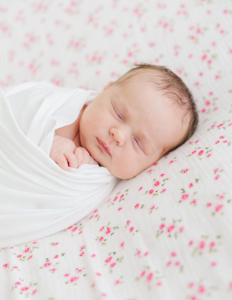 Sweet newborn girl posed on a pink floral backdrop in a bright studio session with an Apex newborn photographer.