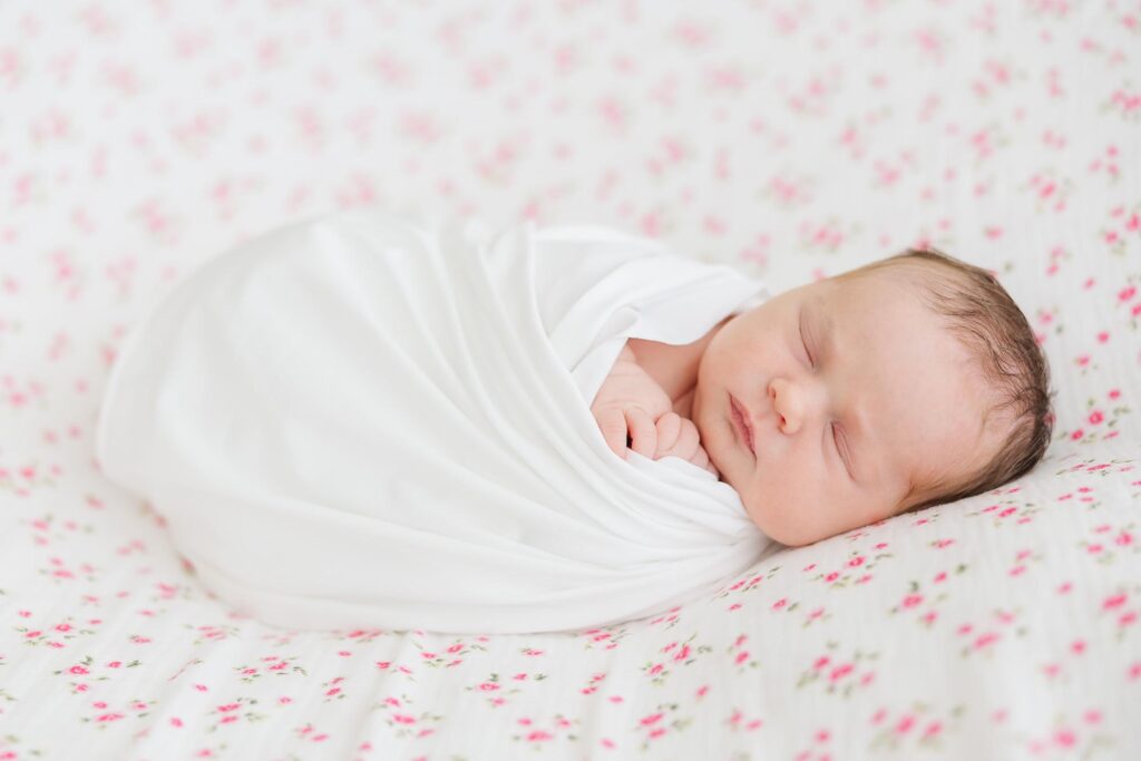 Side profile of a peacefully sleeping newborn wrapped in white on a floral blanket, captured by an Apex newborn photographer.