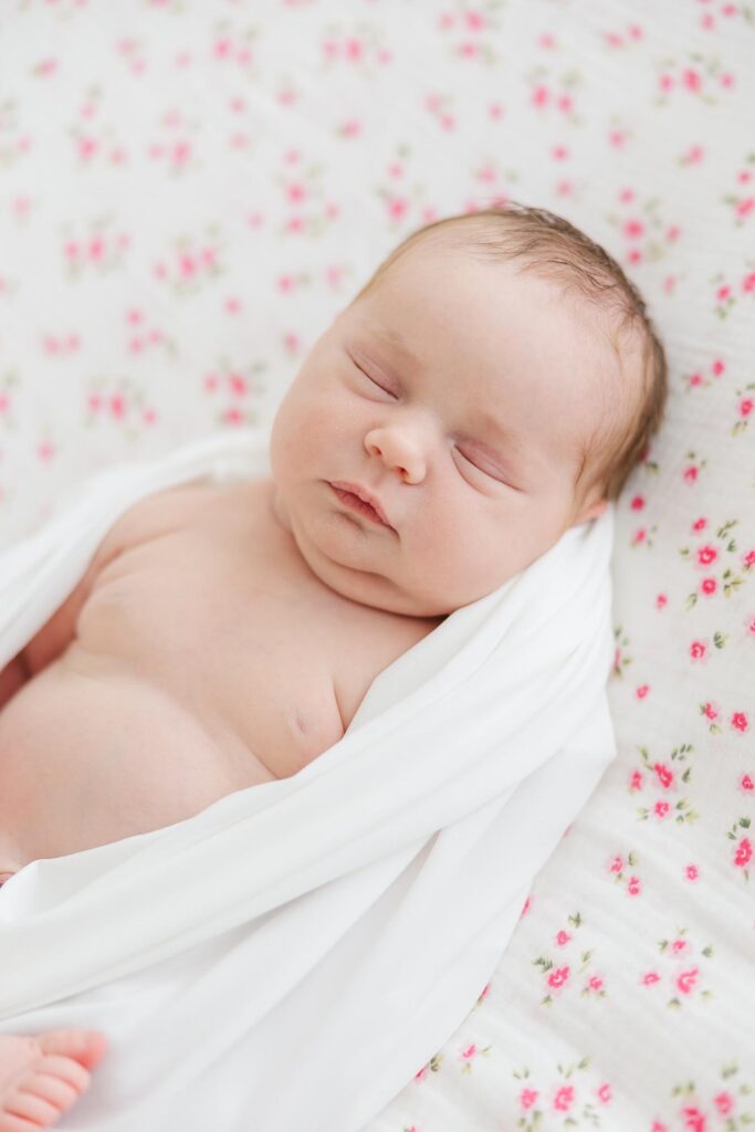 Newborn baby girl swaddled in white on a soft pink floral backdrop during an Apex newborn photographer session.