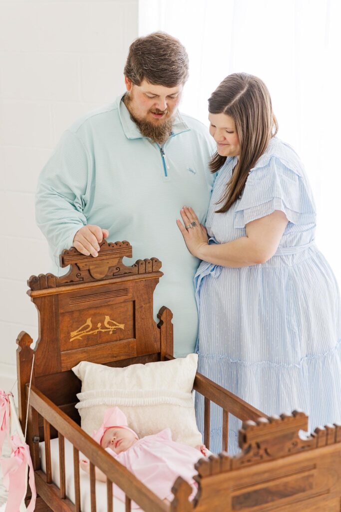 New parents standing beside a wooden heirloom cradle with their newborn daughter during a studio session with an Apex newborn photographer.