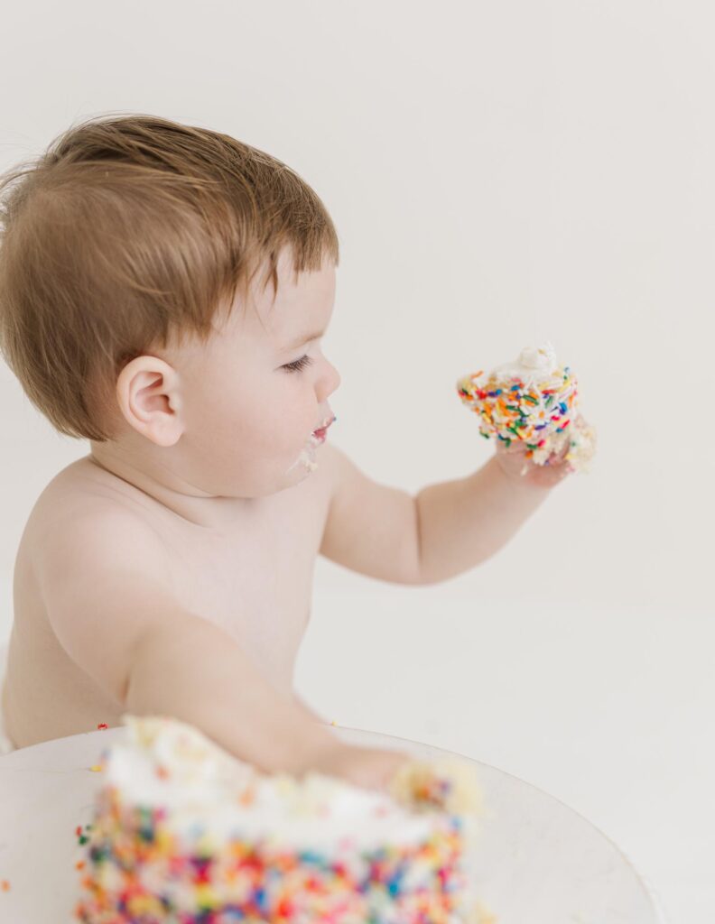 Baby holding piece of sprinkle cake during first birthday cake smash photography session | Raleigh cake smash photographer