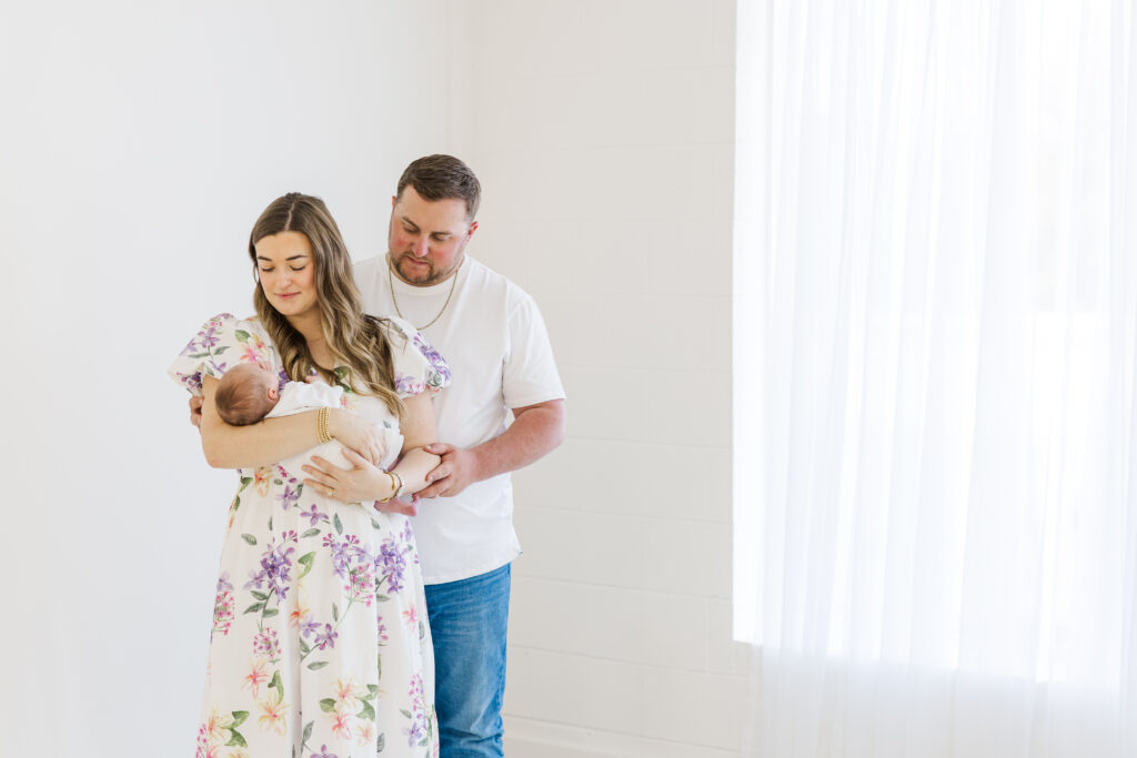 Mother in white dress with pastel florals holding newborn daughter with husband behind her taken by a raleigh studio newborn photographer